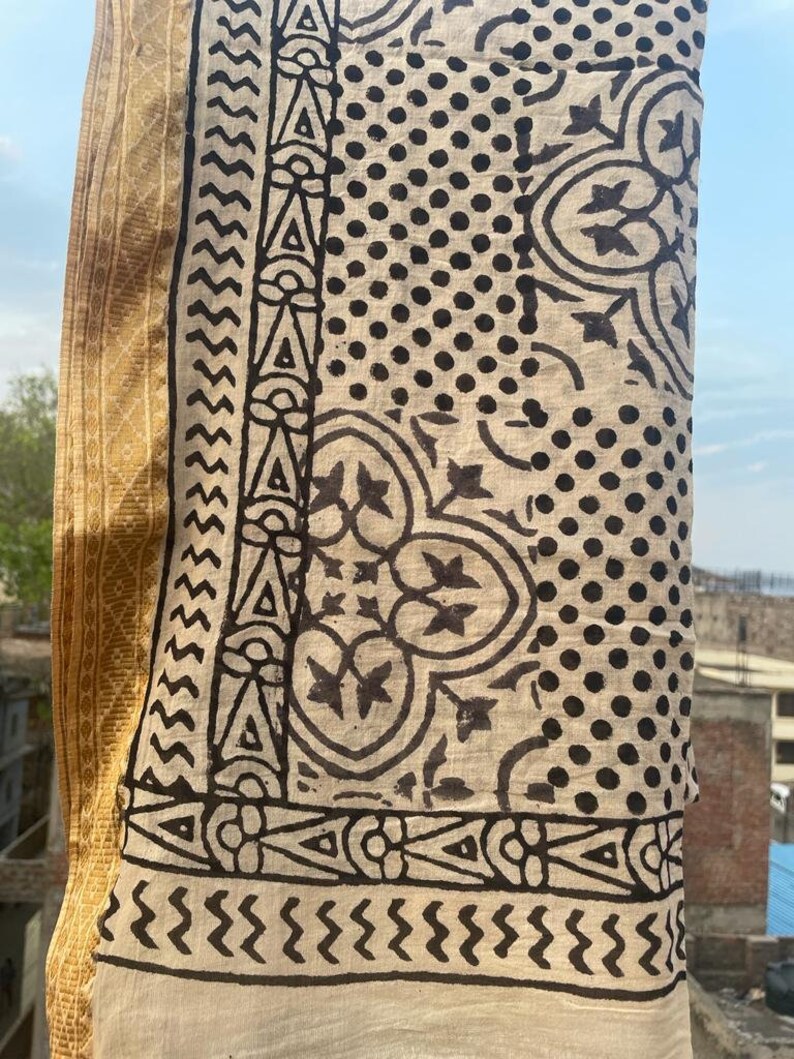 May include: A beige cotton fabric with a black and white block print design. The fabric features a geometric pattern with dots, zigzags, and floral motifs.