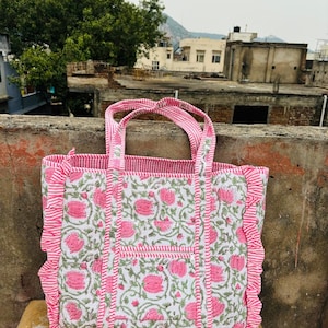 May include: A pink and white tote bag with a floral print and ruffled edges. The bag has two handles and a striped design.
