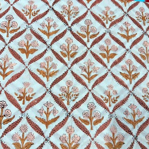 Op de afbeelding: Witte stof met een herhalend patroon van roze bloemen en bruine stelen en bladeren, geplaatst in een ruitvormig raster van bruine lijnen. Het bloemmotief is in blokdrukstijl, wat een textuur geeft.