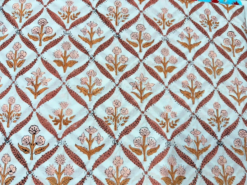 Op de afbeelding: Witte stof met een herhalend patroon van roze bloemen en bruine stelen en bladeren, geplaatst in een ruitvormig raster van bruine lijnen. Het bloemmotief is in blokdrukstijl, wat een textuur geeft.