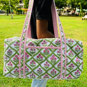 Puede incluir: Bolso tote rectangular grande con fondo blanco y un estampado floral repetido en rosa y verde. El bolso tiene asas y ribetes a rayas rosas y blancas. El bolso se lleva al aire libre.