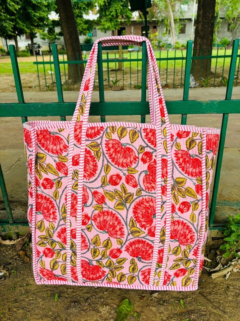 May include: A pink tote bag with a floral print. The bag has white and red stripes around the edges and a pink and white striped handle.