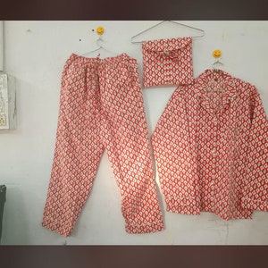 May include: A red and white patterned pajama set, including pants, a button-down shirt, and a matching pouch. The fabric features a repeating diamond design. The set is hanging on a white wall.