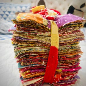 May include: A tall, vertical stack of square fabric swatches in various colours including purple, orange, red, and yellow. The stack is bound together by a single strip of yellow and red fabric tied at the top. Each swatch features white running stitches.
