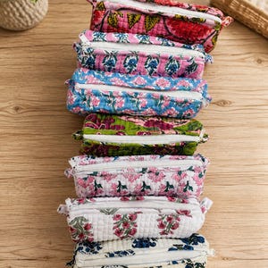 Op de afbeelding: Een stapel van zeven rechthoekige, doorgestikte etuis van stof, elk voorzien van een witte ritssluiting en verschillende florale blokprintpatronen in diverse kleuren zoals roze, blauw, groen en wit, verticaal gerangschikt op een houten ondergrond.
