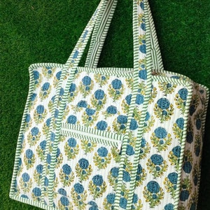 Puede incluir: Una bolsa de mano blanca con un estampado floral verde y azul. La bolsa tiene dos asas y un bolsillo con cremallera en la parte delantera.