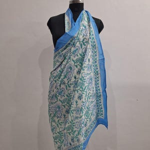 May include: A rectangular scarf featuring a white base with a repeating floral and leaf pattern in shades of blue and green. The scarf has a solid light blue border along the edges and is draped over a black mannequin torso for display.