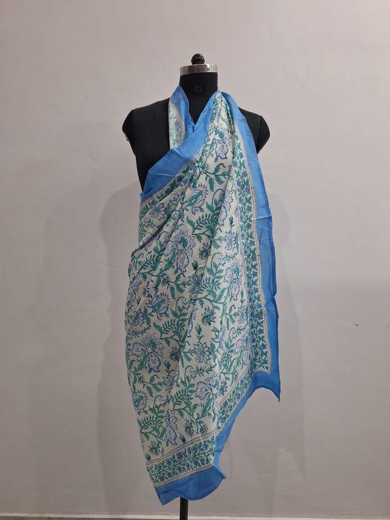 May include: A rectangular scarf featuring a white base with a repeating floral and leaf pattern in shades of blue and green. The scarf has a solid light blue border along the edges and is draped over a black mannequin torso for display.