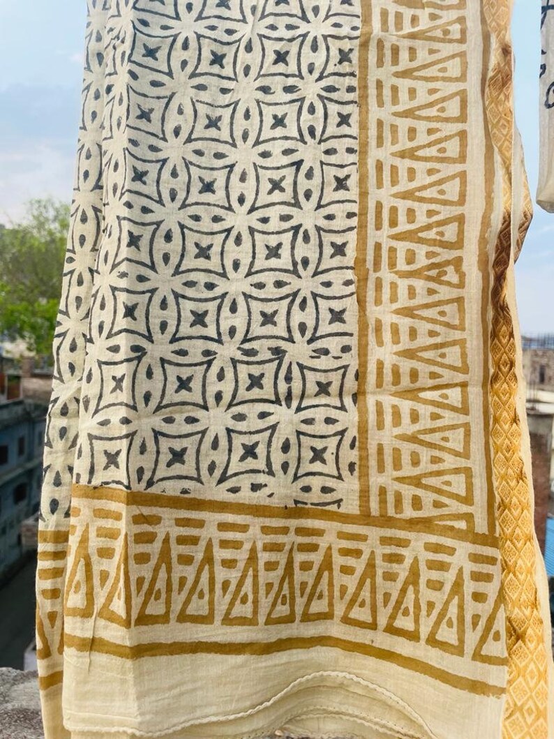 May include: A white cotton scarf with a black and brown geometric pattern. The scarf has a border of brown triangles.