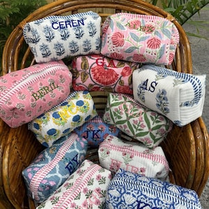 May include: A collection of quilted cosmetic bags in various floral patterns and colors, including pink, blue, and white. Each bag features embroidered initials or names, and is arranged in a wicker chair.