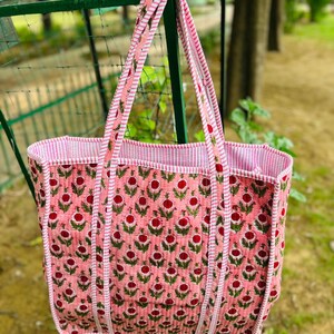 May include: A pink and white quilted tote bag with a floral print. The bag has two long straps and a wide base.