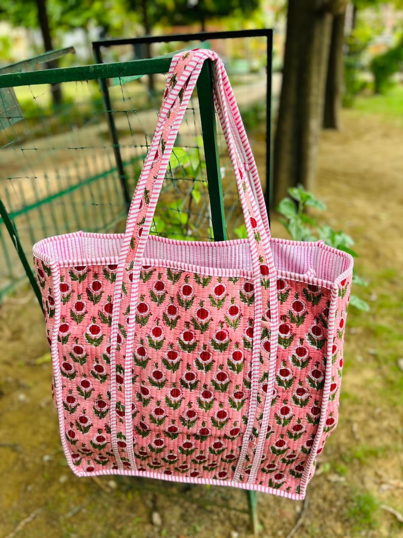 May include: A pink and white quilted tote bag with a floral print. The bag has two long straps and a wide base.