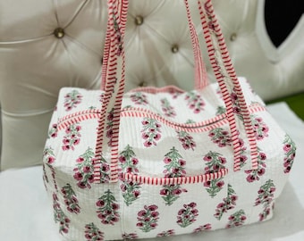 May include: White tote bag with a floral pattern in pink and green. The bag has red striped trim and matching straps. The bag is sitting on a white surface.