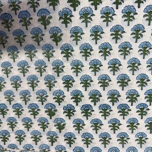 Op de afbeelding: Witte stof met een herhalend patroon van blauwe en groene bloemmotieven. De bloemen hebben een gestileerde uitstraling, met afgeronde bloemblaadjes en gedetailleerde groene stelen en bladeren. Het algehele effect is een delicaat en ingewikkeld patroon.