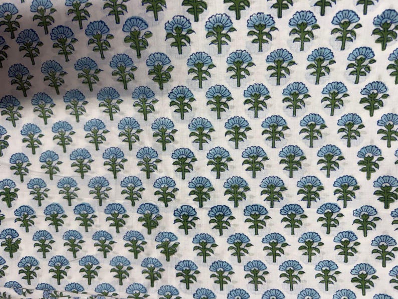 Op de afbeelding: Witte stof met een herhalend patroon van blauwe en groene bloemmotieven. De bloemen hebben een gestileerde uitstraling, met afgeronde bloemblaadjes en gedetailleerde groene stelen en bladeren. Het algehele effect is een delicaat en ingewikkeld patroon.