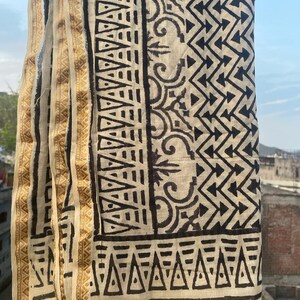 May include: A white cotton scarf with a black geometric pattern. The scarf has a gold border with a repeating pattern.