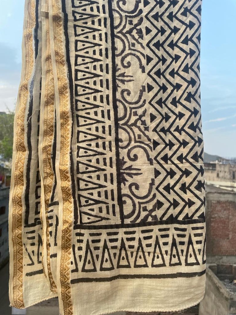 May include: A white cotton scarf with a black geometric pattern. The scarf has a gold border with a repeating pattern.