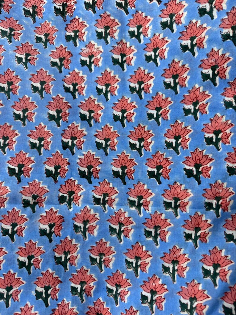 Op de afbeelding: Een lichtblauwe stof met een herhalend patroon van roze lotusbloemen en groene bladeren. Het bloemmotief is gelijkmatig verdeeld over het textiel, waardoor een symmetrische opstelling ontstaat. De stof lijkt van katoen of een vergelijkbaar materiaal te zijn.