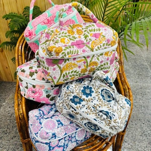 May include: A collection of colorful, patterned cosmetic bags with zippered closures and top handles. The bags feature floral and geometric designs in pink, blue, and white, and are arranged on a wicker chair.