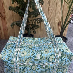 May include: A large, rectangular tote bag with a floral pattern in shades of blue, green, and white. The bag has two long handles with a striped design. The bag is made of a quilted fabric.