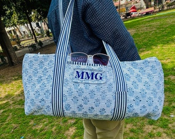 May include: A light blue and white floral print tote bag with striped handles and a front pocket. The pocket features the embroidered initials "MMG". Sunglasses rest in the pocket. The bag is approximately 20 inches long.