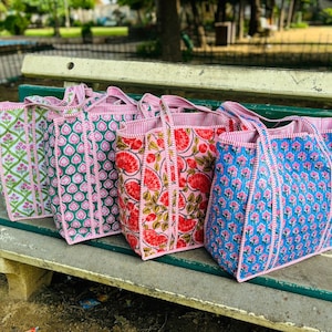 May include: Four colorful tote bags with pink trim and floral patterns. The bags are lined up on a wooden bench.