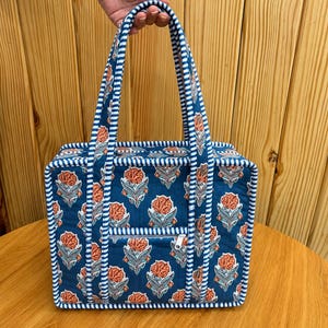 May include: A rectangular quilted tote bag featuring a dark blue fabric with a repeating orange and light blue floral pattern. The bag includes two long handles and a front zippered pocket, all accented with a white and blue striped fabric trim.