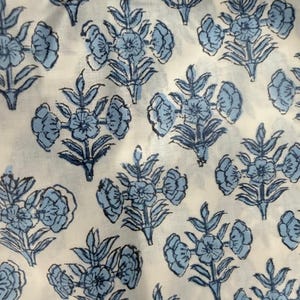 Op de afbeelding: Close-up van een witte stof met een herhalend patroon van blauwe bloemmotieven. De bloemen hebben meerdere bloemblaadjes en zijn gerangschikt in clusters met bladeren. De stof lijkt katoen of linnen te zijn.