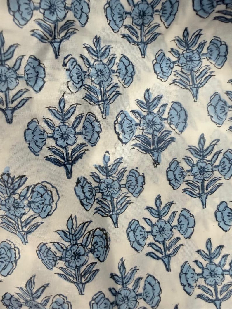 Op de afbeelding: Close-up van een witte stof met een herhalend patroon van blauwe bloemmotieven. De bloemen hebben meerdere bloemblaadjes en zijn gerangschikt in clusters met bladeren. De stof lijkt katoen of linnen te zijn.