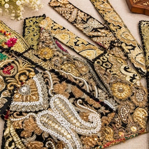 May include: A collection of black fabric strips featuring intricate gold embroidery, metallic sequins, and white pearl bead accents. The decorative borders display ornate floral and paisley patterns with dense stitching and shimmering textures.