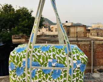 May include: A floral print duffel bag with blue flowers and green leaves on a white background. The bag has a rectangular shape with a pocket on the front and two handles. The handles are striped with blue and white.