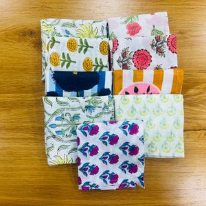 Op de afbeelding: Een verzameling gevouwen stoffen vierkanten met diverse bloemen- en geometrische patronen. Het kleurenpalet omvat wit, geel, roze, blauw en oranje. De stoffen lijken van katoen te zijn, geschikt voor zakdoeken of kleine doeken.