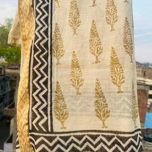 May include: A white cotton scarf with a gold tree print and black zigzag border. The scarf is folded and draped over a surface.