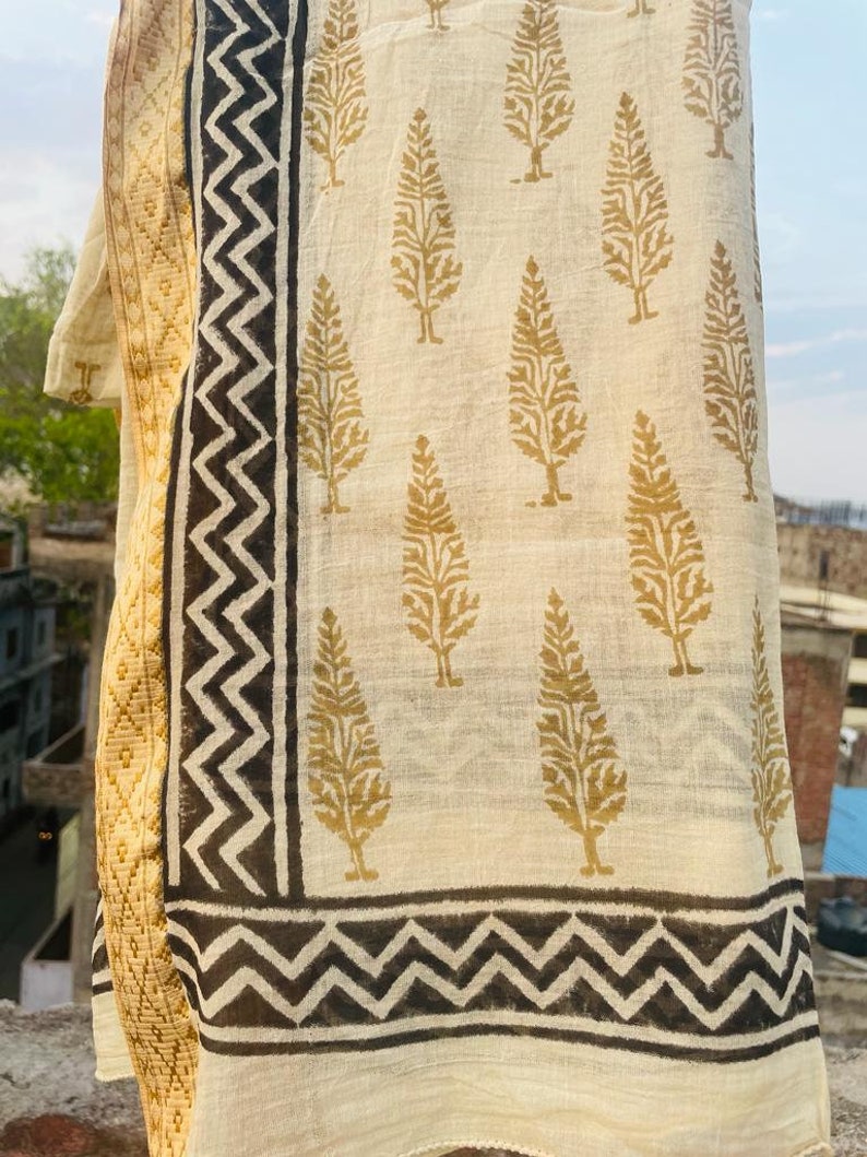 May include: A white cotton scarf with a gold tree print and black zigzag border. The scarf is folded and draped over a surface.