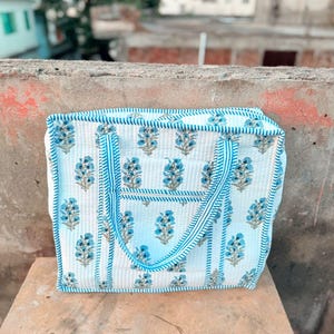 May include: A white quilted tote bag with a blue floral pattern and blue striped trim. The bag has two handles and a rectangular shape. The bag is sitting on a wooden surface.