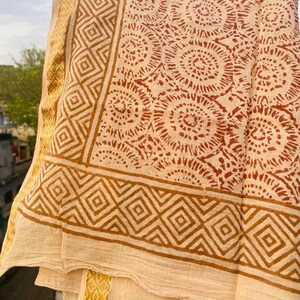 May include: A cream-colored cotton scarf with a brown and orange geometric pattern. The scarf has a border of small diamonds and a larger border of a repeating floral design.