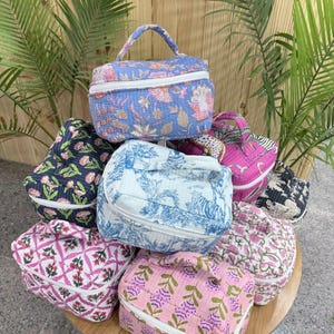 Pode incluir: Uma coleção de nécessaires acolchoadas com vários designs florais e estampados. As bolsas apresentam fecho de correr e uma pega superior. As cores incluem azul, rosa, branco e preto. As bolsas estão dispostas numa mesa de madeira.