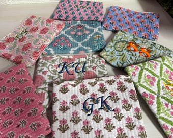 May include: A collection of quilted cosmetic bags in various floral patterns and colors, including pink, blue, and green. Several bags feature embroidered initials in blue or orange thread, adding a personalized touch to the accessories.