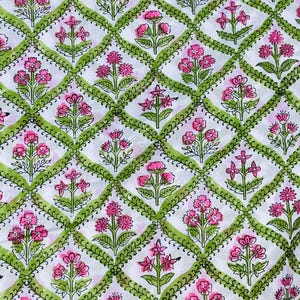 Op de afbeelding: Witte stof met een herhaald patroon van roze bloemen en groene ruitvormige randen. Het bloemmotief bevat verschillende bloemvormen en bladdetails, waardoor een levendig en gedetailleerd textielpatroon ontstaat.