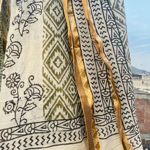 May include: A white cotton fabric with a green and black geometric pattern and floral designs. The fabric has a gold trim along the edge.