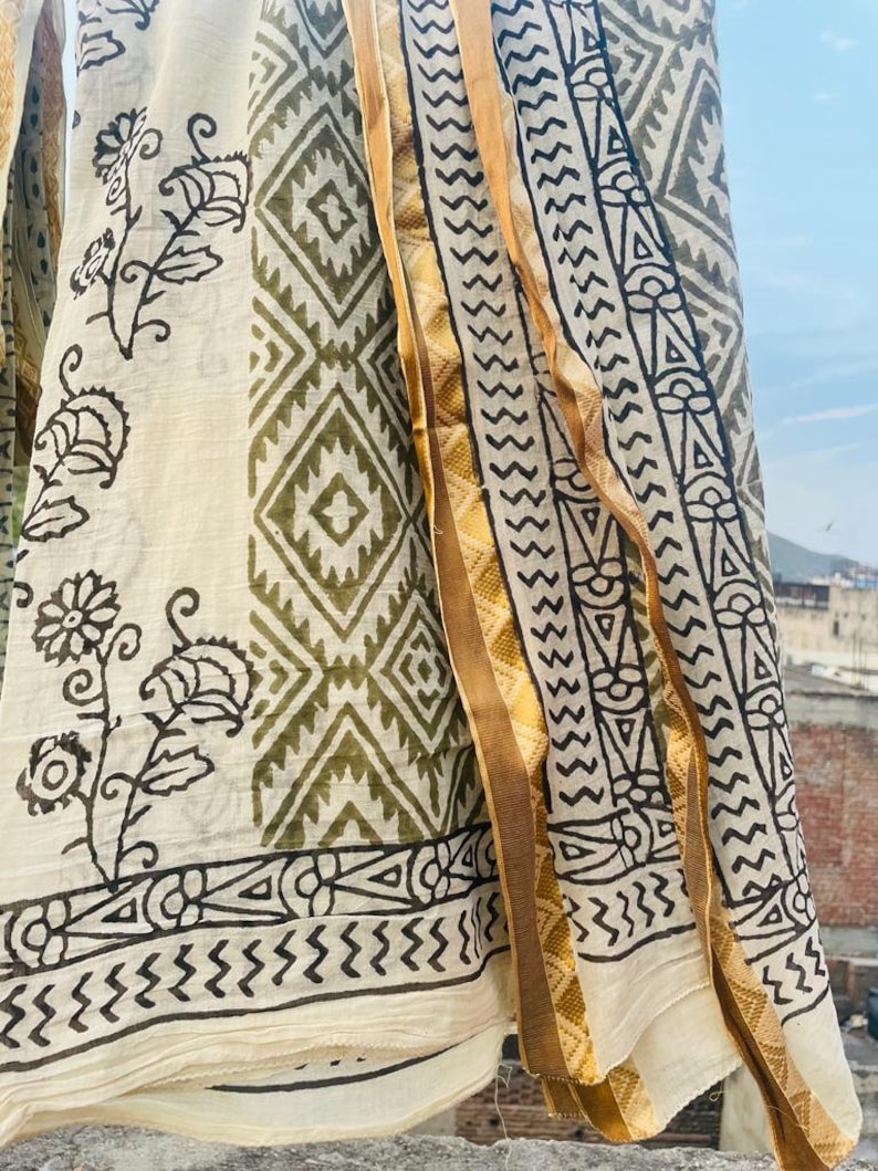 May include: A white cotton fabric with a green and black geometric pattern and floral designs. The fabric has a gold trim along the edge.
