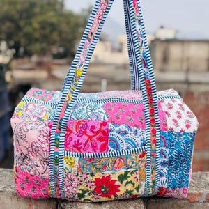 Puede incluir: Un bolso de viaje de patchwork colorido con estampados florales y cachemir en rosa, azul y amarillo. El bolso tiene asas a rayas y forma rectangular. El bolso está hecho de tela acolchada.