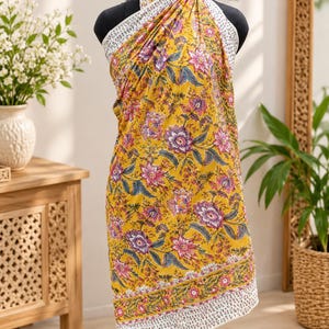 May include: A yellow sarong wrap featuring a vibrant floral pattern in shades of pink, purple, and green. The fabric has a white border with a small, repeating black print along the edges and ties securely around the neck with matching fabric.