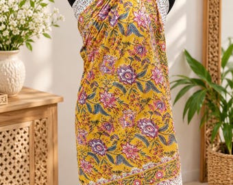 May include: A yellow sarong wrap featuring a vibrant floral pattern in shades of pink, purple, and green. The fabric has a white border with a small, repeating black print along the edges and ties securely around the neck with matching fabric.