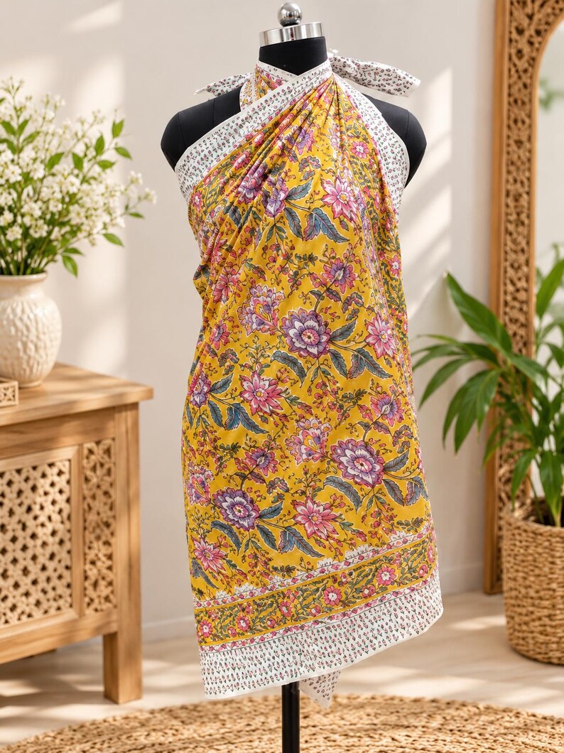 May include: A yellow sarong wrap featuring a vibrant floral pattern in shades of pink, purple, and green. The fabric has a white border with a small, repeating black print along the edges and ties securely around the neck with matching fabric.