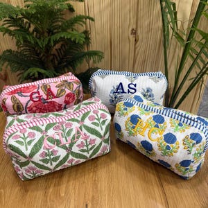 Può includere: Quattro trousse rettangolari con motivi floreali e iniziali ricamate. Una è rosa con fiori rossi e "GA", un'altra è bianca con fiori blu e "AS", una terza è bianca con fiori rosa e "CLA", e l'ultima è bianca con fiori gialli e "HC".