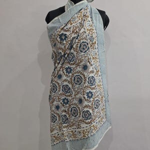 May include: A sarong featuring a white base with a repeating floral pattern in muted blue and brown tones. The fabric includes a border with thin blue and white horizontal stripes along the edges, draped over a black mannequin torso.