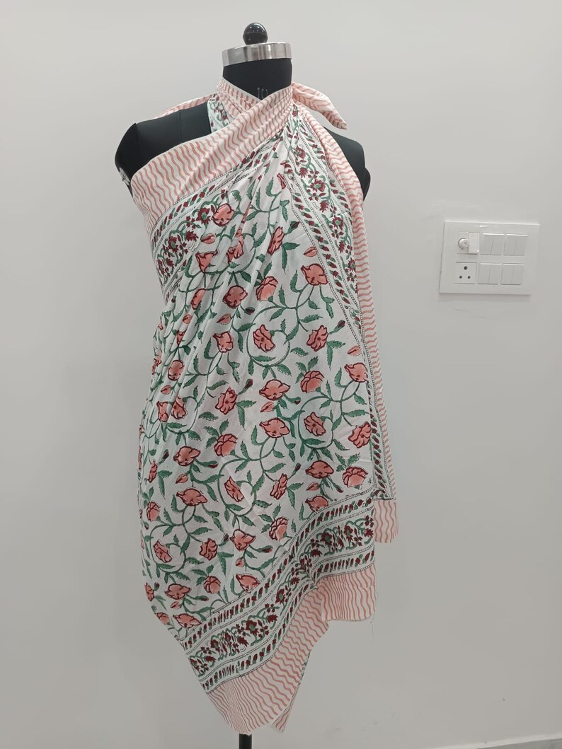 May include: A sarong featuring a white base with a repeating floral pattern in shades of pink and green. The fabric has a border with a pink and white wavy line design. The garment is draped around a mannequin, tied at the neck with a halter style.