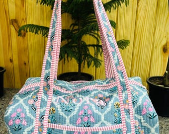 May include: A quilted duffel bag in a light blue fabric featuring a white lattice pattern and pink and green floral motifs. The bag has pink and white striped fabric handles and trim, with the name NORA embroidered in pink on the front pocket.