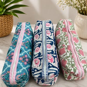 May include: Three rectangular quilted pouches with zippered closures. The left pouch is teal with a pink floral pattern, the center is navy blue with pink flowers, and the right is white with pink floral motifs. Each features a pink and white striped zipper.